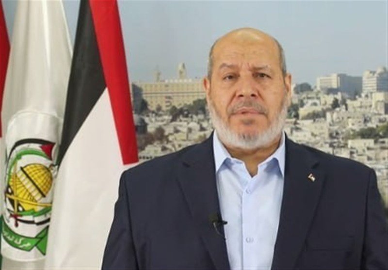 A bald, bearded man in a dark suit and light blue shirt speaks into a lapel microphone, with a flag and cityscape visible in the background.