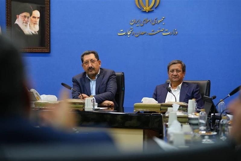 Two men in suits sit at a conference table with microphones, water bottles, and tissues, against a blue wall featuring Persian text, an emblem, and a framed portrait.