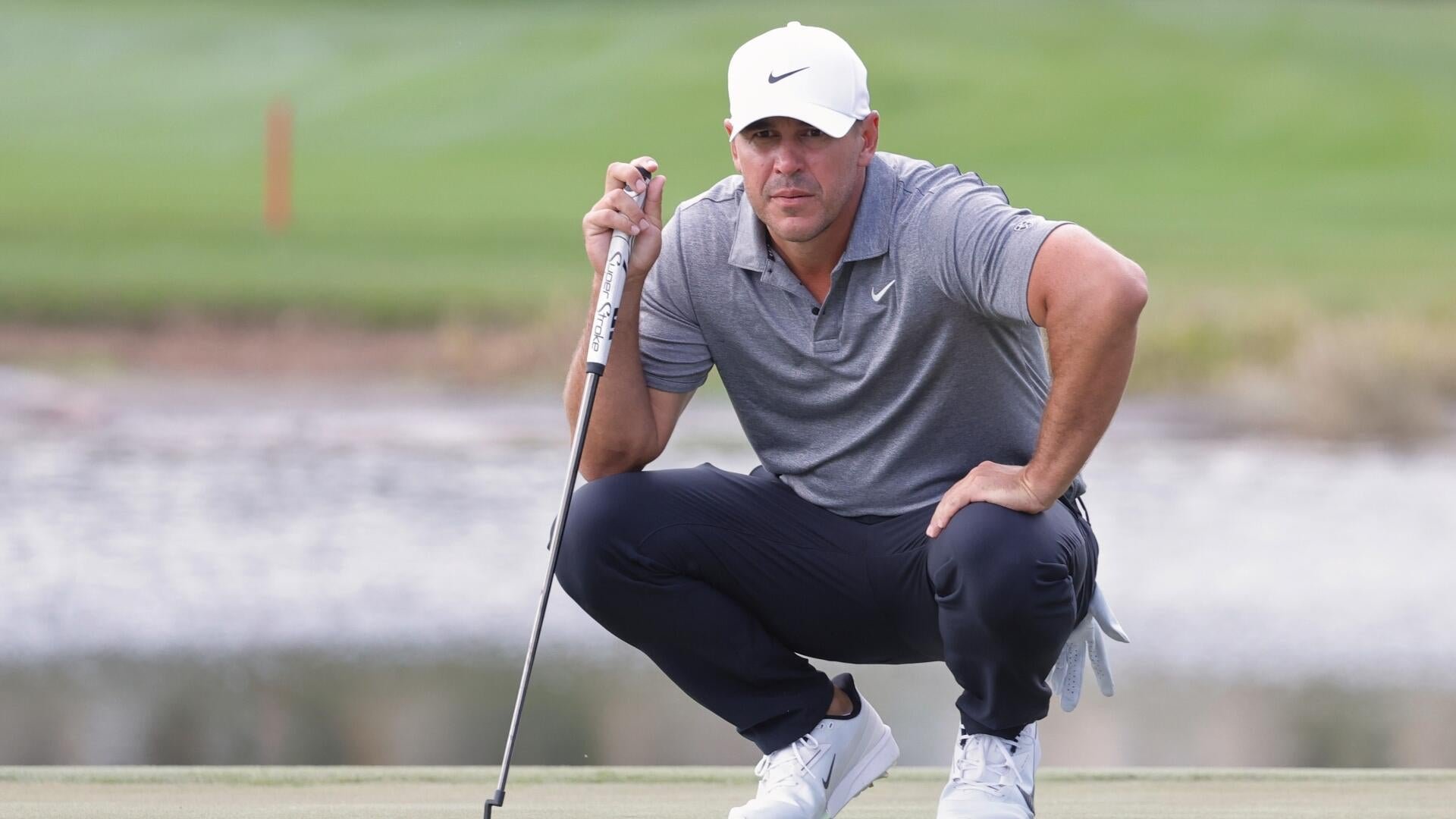 A golfer in a white Nike cap and gray polo shirt crouches on a green, holding a putter while studying a putt.