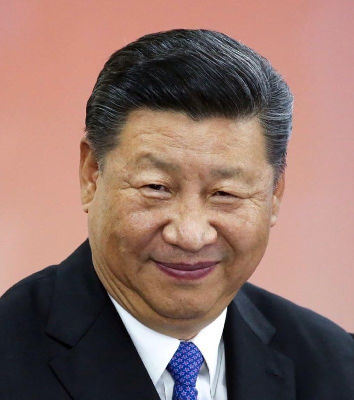 A smiling man with graying hair wears a dark suit, white shirt, and blue tie against a soft pink backdrop.