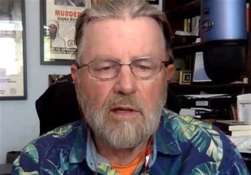 A bearded man with glasses wearing a tropical-patterned shirt speaks in an office setting with framed posters and bookshelves visible behind him.