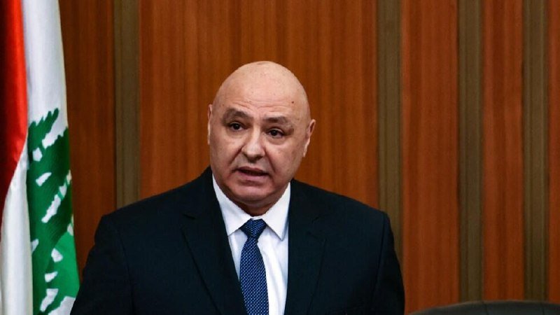 A bald man in a dark suit and blue tie speaks at a podium, with a red, white, and green flag visible behind him.