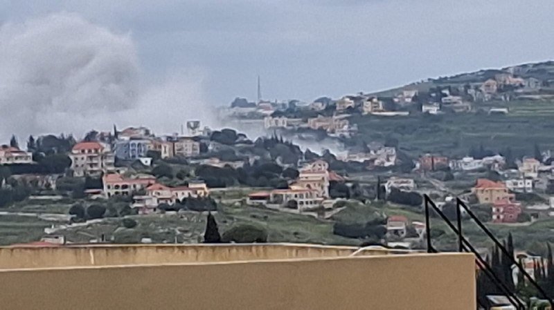 A plume of gray smoke rises from a hillside residential area dotted with houses and greenery under a cloudy sky.