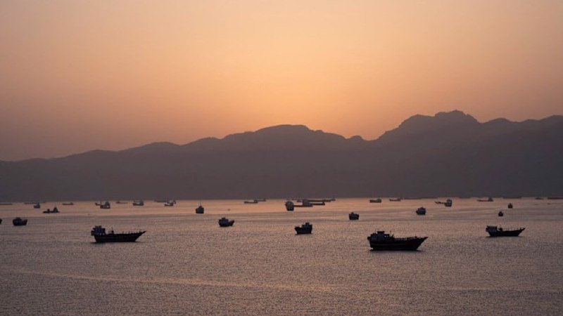 Numerous small boats float on calm waters at dusk, silhouetted against a distant mountain range under a hazy, peach-colored sky.