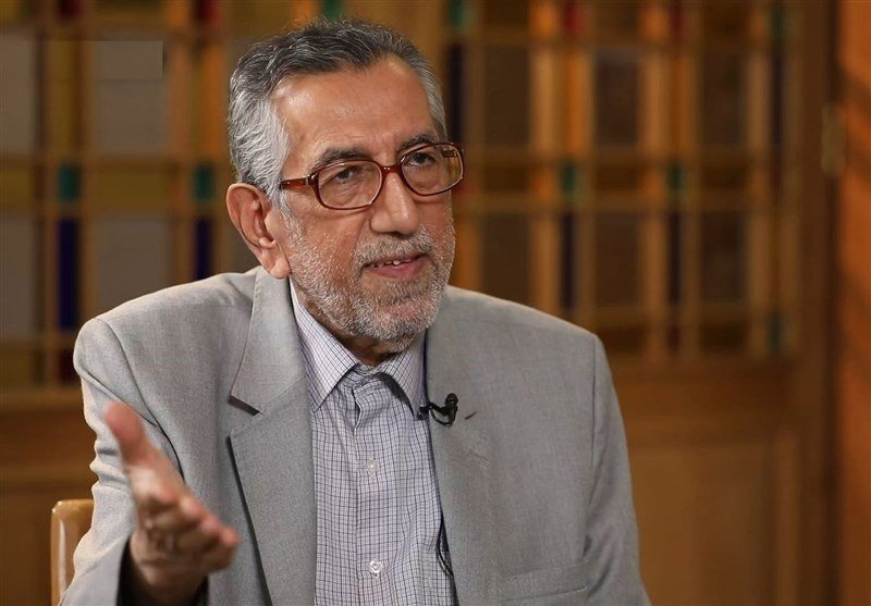 A gray-haired, bearded man wearing glasses and a gray suit gestures with his hand during what appears to be an interview, with a blurred wooden lattice background.