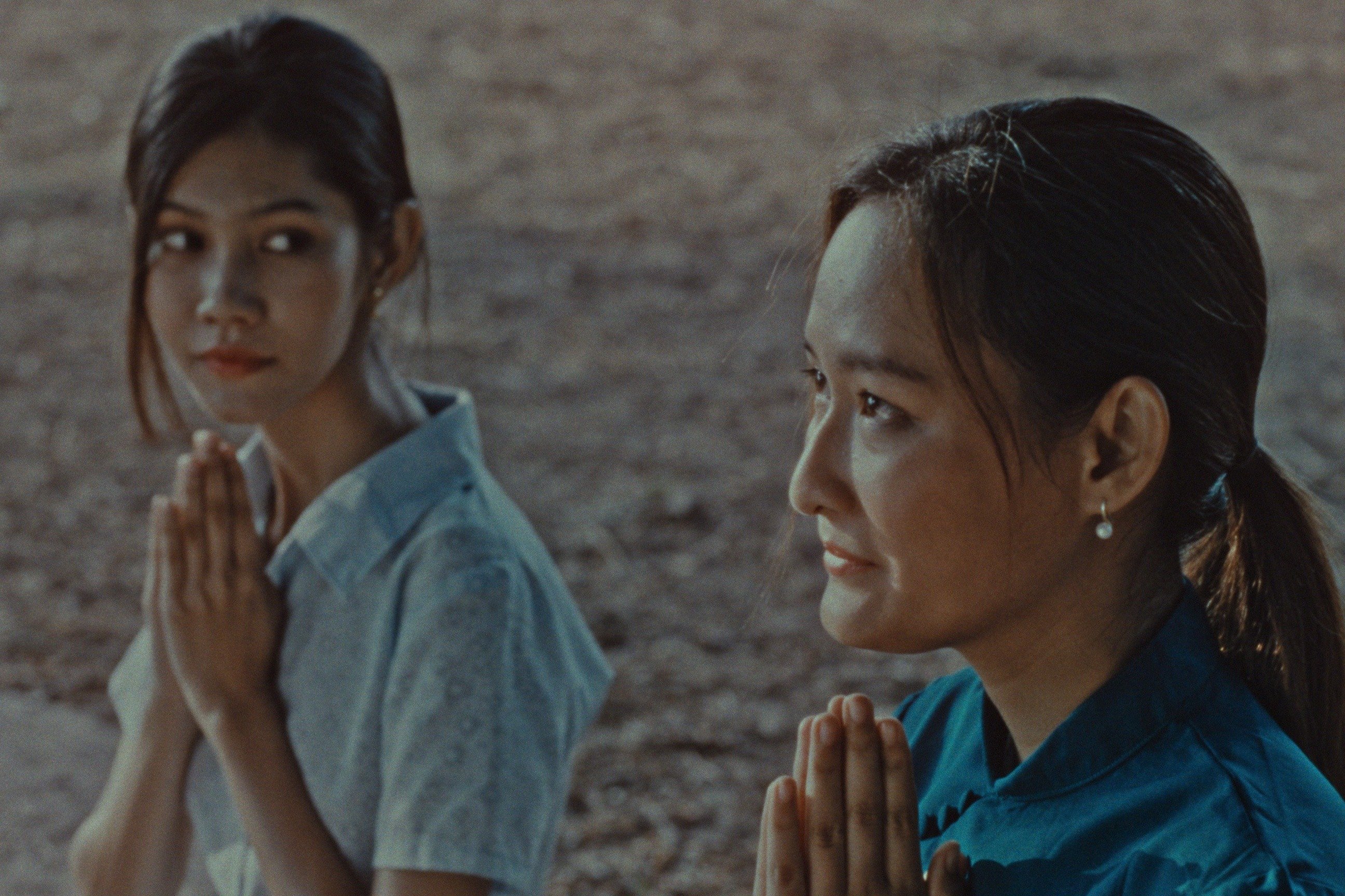 Two women kneel with hands pressed together in a prayer gesture against a whitewashed brick wall.