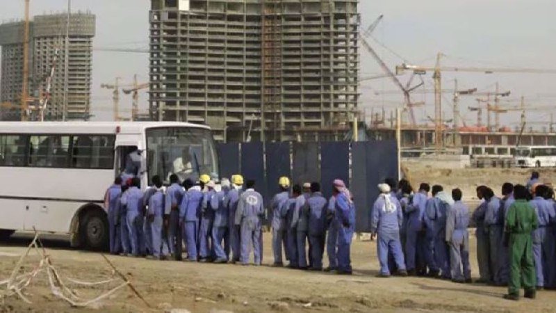 Workers in blue coveralls and hard hats line up to board a white bus at a large construction site with high-rise buildings and multiple cranes in the background.