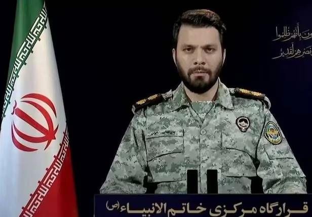 A bearded man in a camouflage military uniform with insignia sits at a podium beside an Iranian flag, with Persian text displayed on the screen.