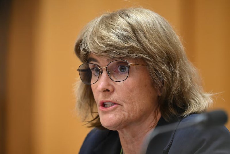 A woman with shoulder-length blonde hair and round wire-rimmed glasses speaks, her mouth slightly open, against a blurred wood-paneled background.