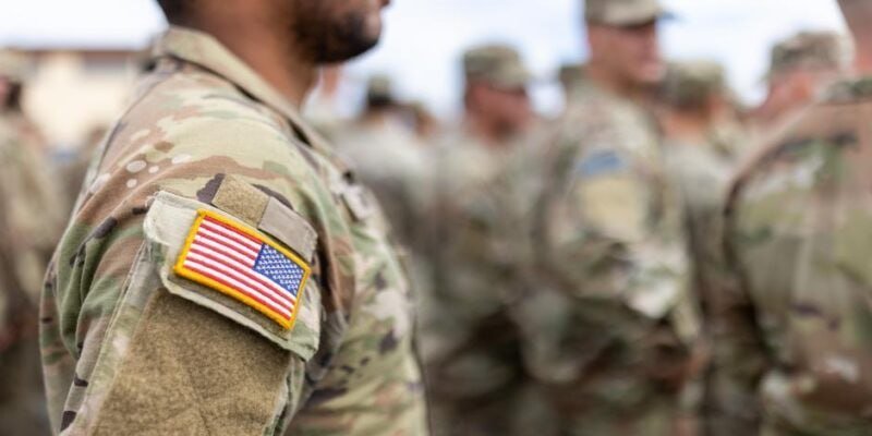 A close-up shows a soldier in camouflage uniform with a U.S. flag patch on the shoulder, standing outdoors among a blurred group of similarly dressed military personnel.
