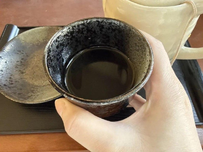 A hand holds a dark ceramic cup containing a clear liquid, with a saucer and pale teapot visible on a black tray.