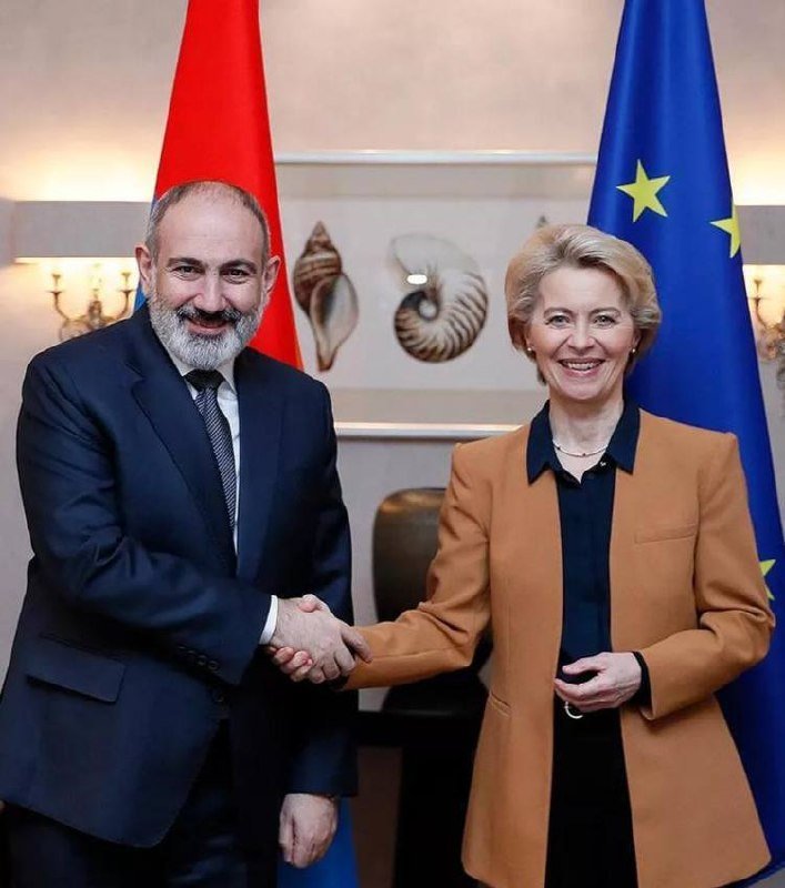A bearded man in a dark suit and a blonde woman in a tan blazer shake hands, smiling, with red, blue, and EU flags visible behind them.