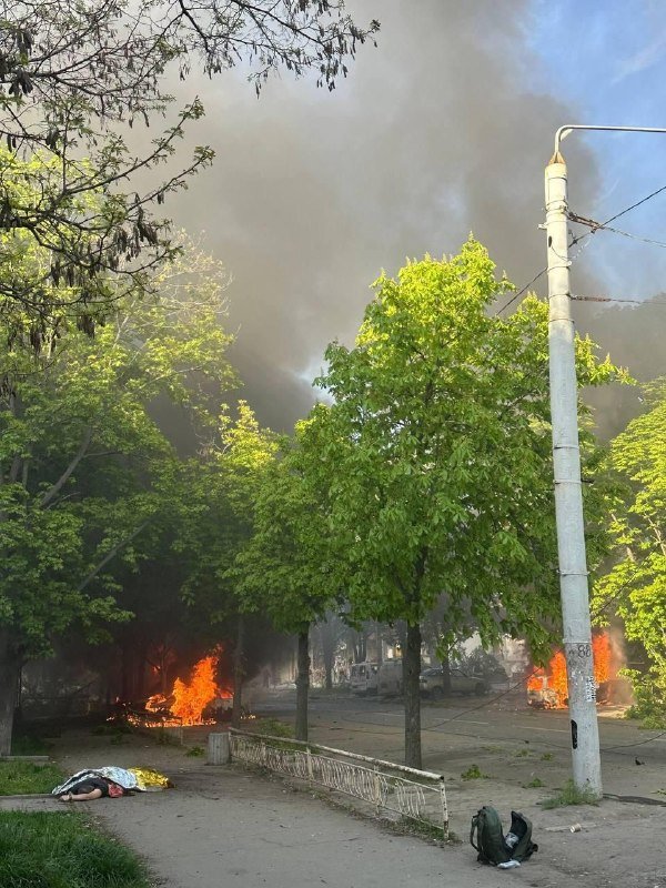 A large fire burns along a tree-lined street, sending thick black smoke into the sky, while a person lies motionless on the pavement near a fallen fence and backpack.