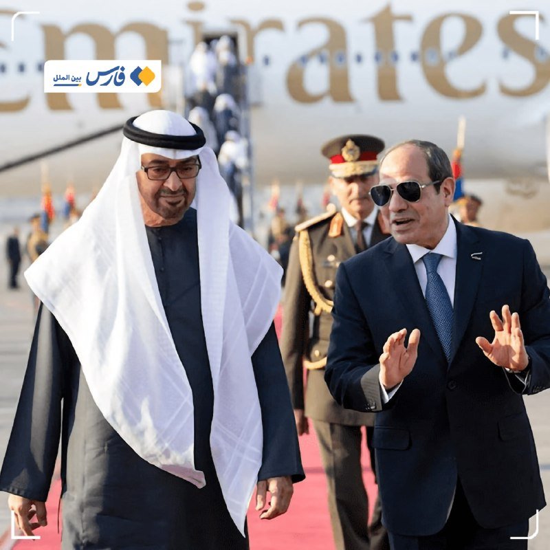Two men in formal attire—one in traditional Gulf dress, the other in a dark suit and sunglasses—walk along a red carpet past a military officer, with an Emirates airplane visible behind them.