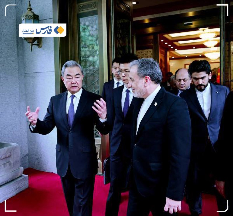 A group of men in dark suits walk together through an ornate, well-lit hallway with red carpeting and decorative wall lanterns.