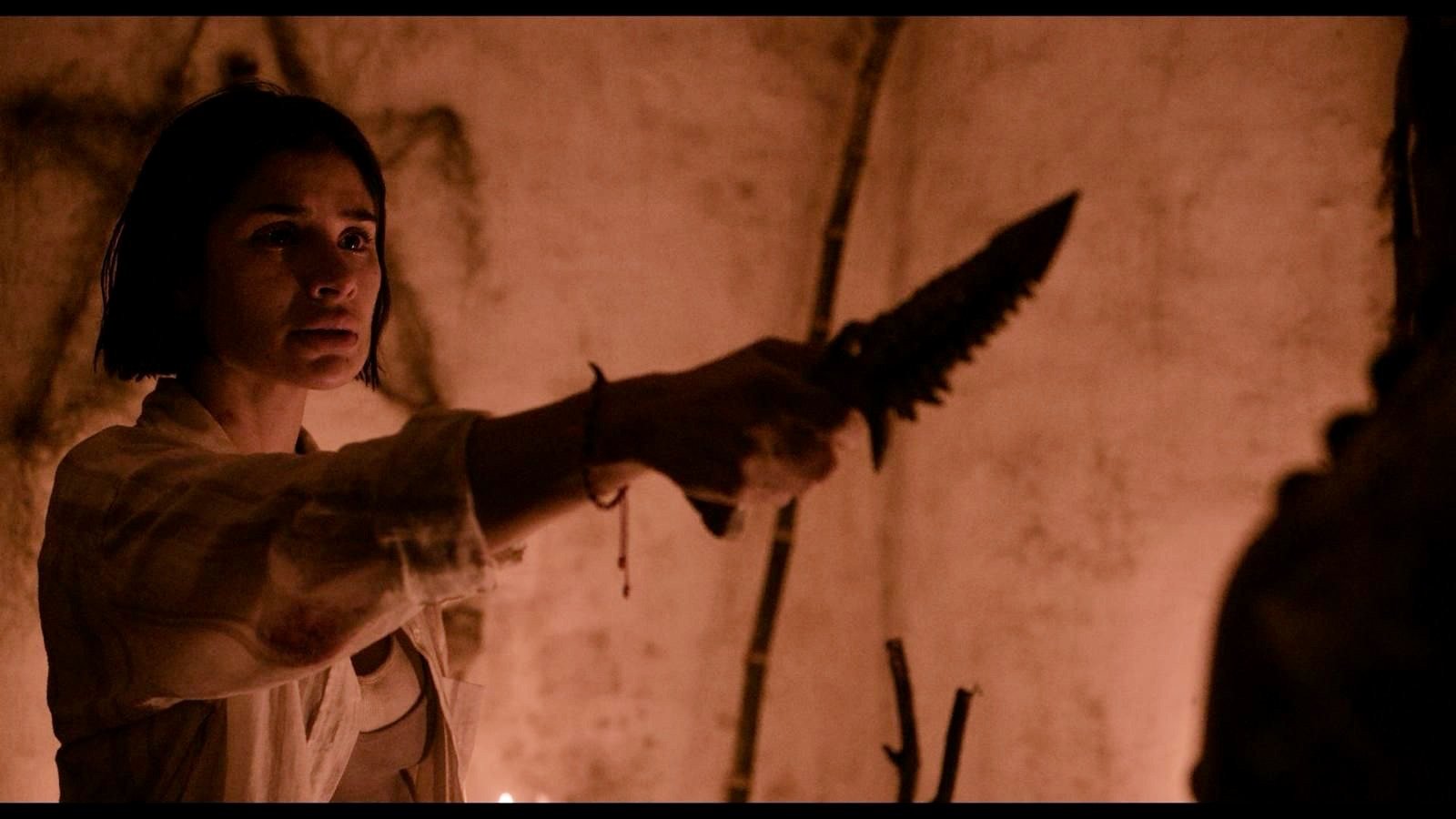 A woman with short dark hair holds a serrated knife forward in a dimly lit, reddish-toned interior with cracked walls.