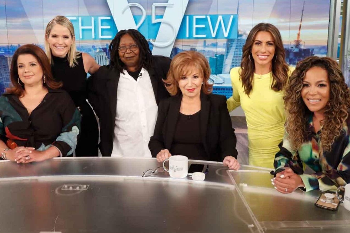 Six women pose together at a studio desk in front of a "The View" logo backdrop with a city skyline.