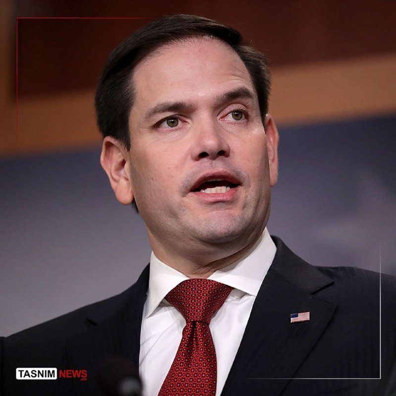 A man in a dark suit, white shirt, and red patterned tie speaks while wearing an American flag lapel pin, with a "Tasnim News" watermark visible.