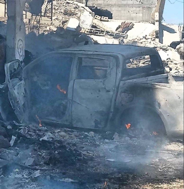 A burned-out SUV with an open door sits amid rubble and concrete debris, with smoke and small flames still visible near the vehicle.