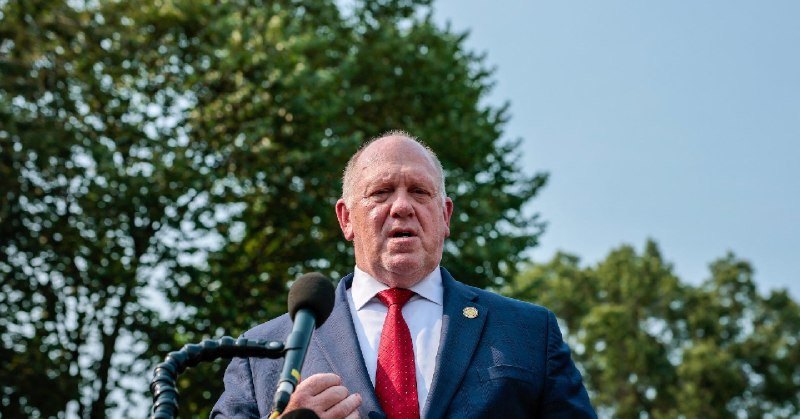 A man in a navy suit and red tie speaks into a microphone outdoors with trees in the background.