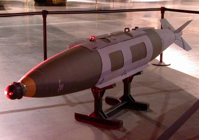 A cylindrical, gray and white aerial bomb with rear fins is displayed on a black stand behind a stanchion barrier.