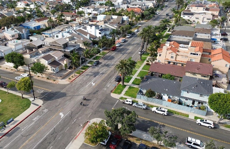 An aerial view shows a residential intersection lined with palm trees, parked cars, and houses with varied rooflines on a sunny day.
