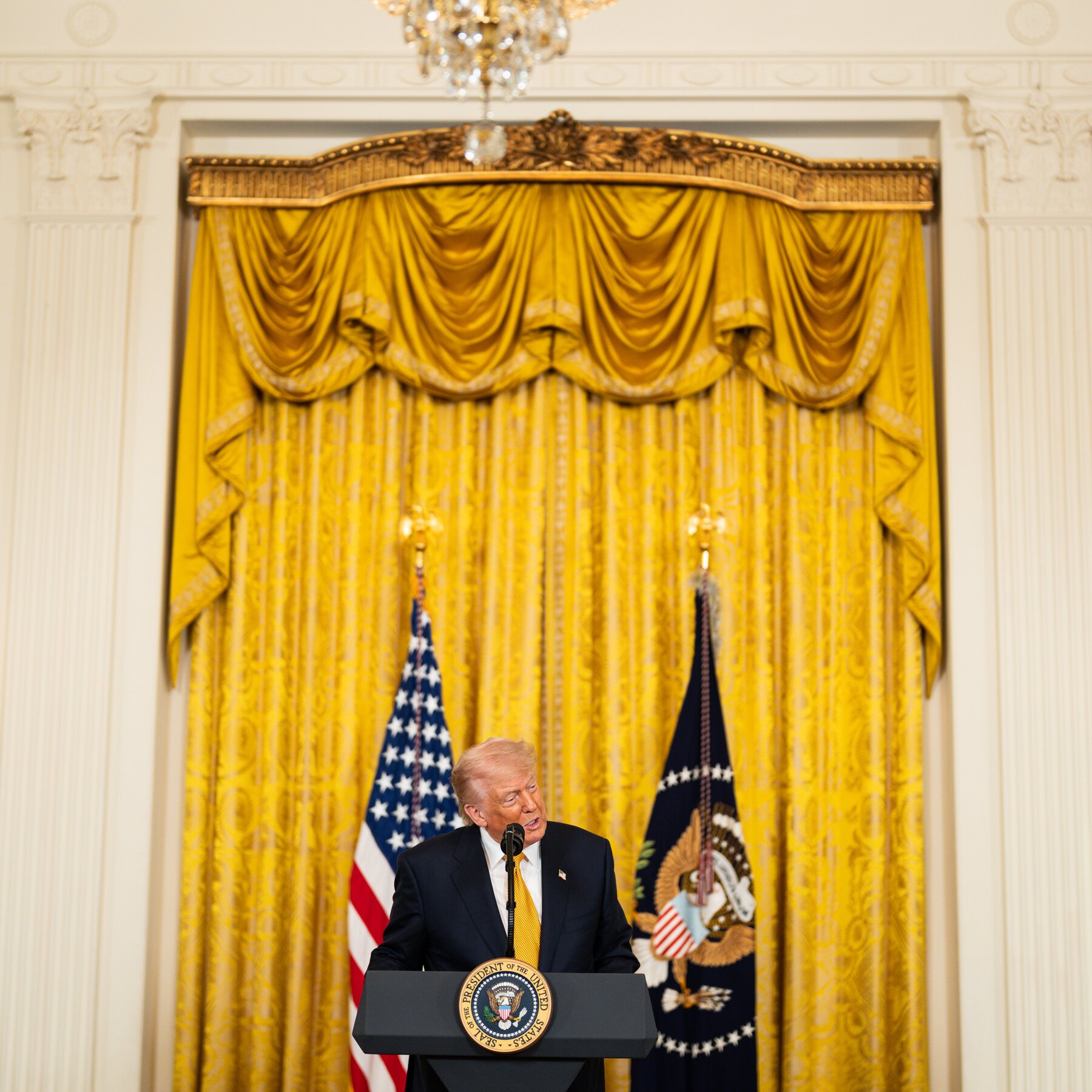A man in a dark suit and yellow tie speaks at a podium bearing the presidential seal, flanked by the American flag and another flag against golden drapes.