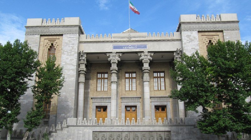 A stone building with tall columns, arched doorways, and crenellated towers stands beneath a flag of Iran.