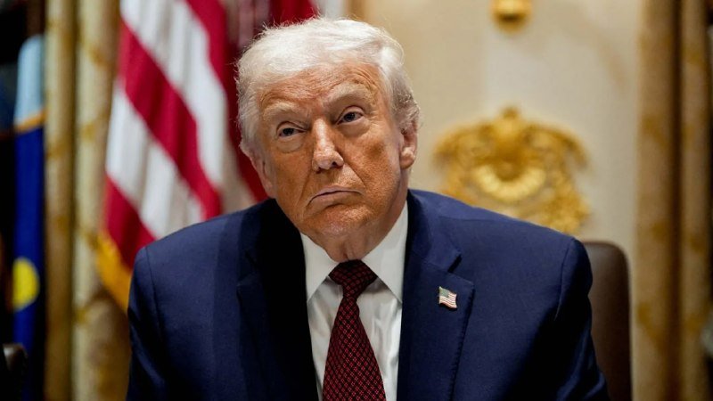 A white-haired man in a dark suit and red patterned tie sits before an American flag and an ornate gold-trimmed background, looking off-camera with a serious expression.