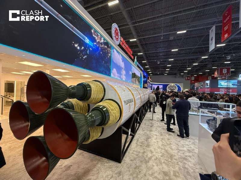 A large rocket model labeled "MIDCLASOR" is displayed at an indoor trade show, with attendees gathered around viewing exhibits under a high industrial ceiling.