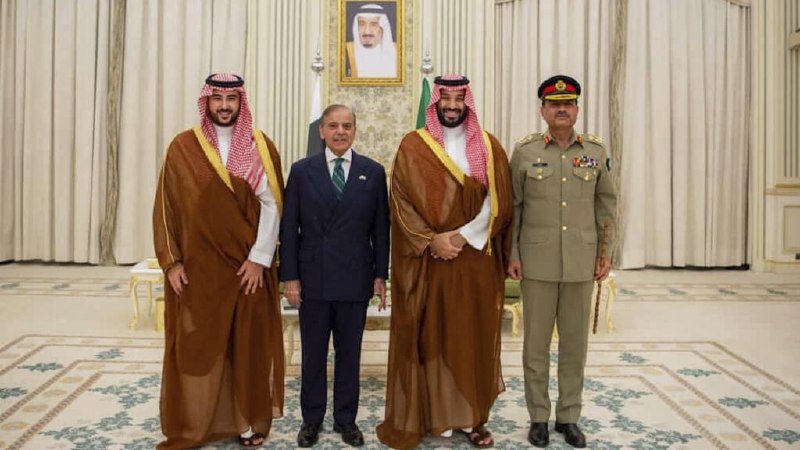 Four men, including two in traditional Arab robes, one in a dark suit, and one in a military uniform, stand together indoors beneath a framed portrait.