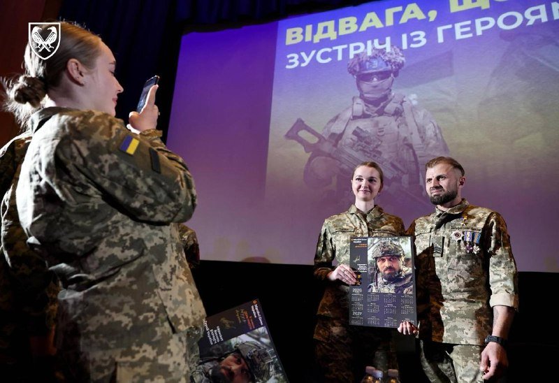 Two soldiers in camouflage uniforms pose on stage holding a calendar featuring a soldier's portrait, while another servicewoman in the foreground photographs them with her phone beneath a projected image of an armed soldier and Ukrainian text.