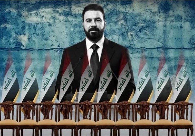 A bearded man in a dark suit stands behind a row of empty chairs, with multiple Iraqi flags displayed in the foreground against a textured blue backdrop.