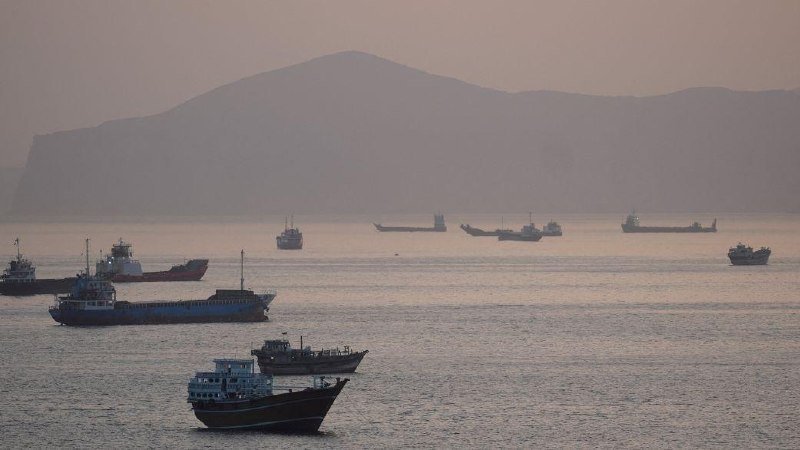 Cargo ships and small boats float on calm waters near a hazy mountain coastline at dusk.