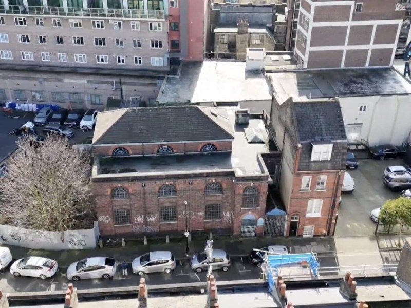 An aerial view shows a brick building with arched windows beside a parking area where several cars are parked along a graffiti-marked wall.