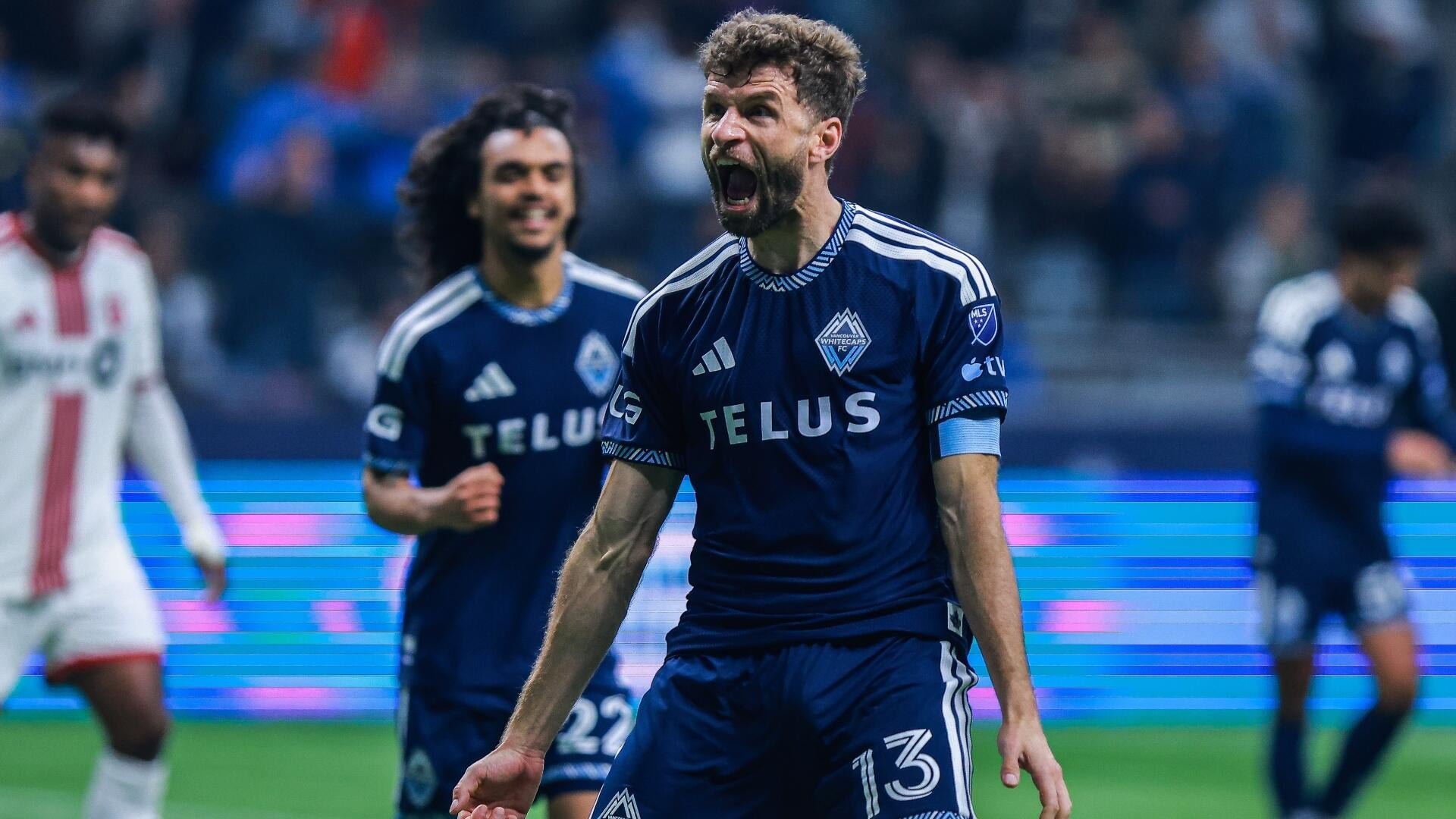 A Vancouver Whitecaps FC player wearing a navy "TELUS" jersey with number 15 screams in celebration during an MLS match, with teammates and a crowd visible behind him.