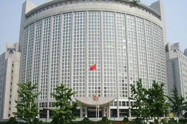 A large, curved, multi-story government building with extensive glass windows features a red flag flying at its central entrance, with green trees lining the foreground.