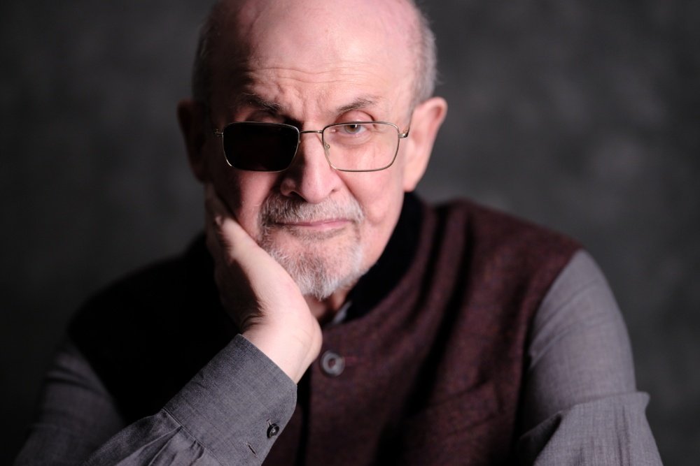 A balding older man with a white beard, wearing glasses and a dark eye patch over his right eye, rests his chin on his hand against a dark gray background.