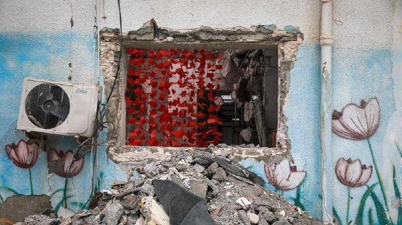 A damaged building wall with a broken window opening revealing a red perforated interior panel, flanked by an air conditioning unit and floral murals amid rubble.