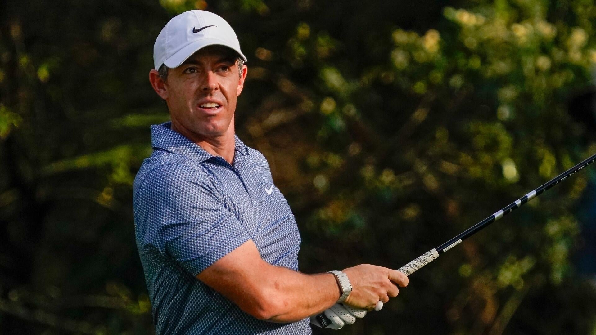 A male golfer in a white Nike cap and patterned blue polo shirt holds a golf club during a swing, with green trees in the background.