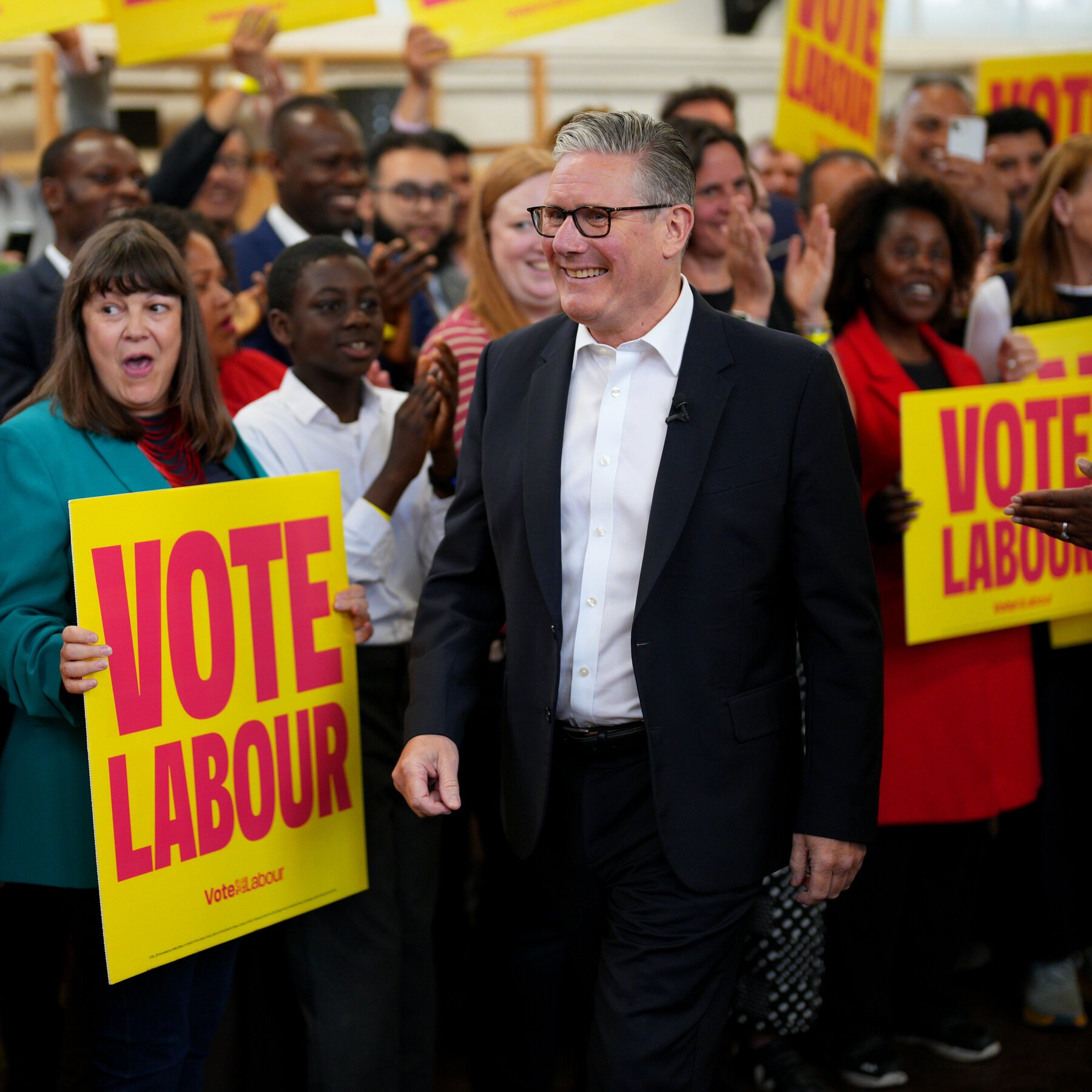 A smiling man in a dark suit and glasses walks through a crowd of supporters holding yellow "Vote Labour" signs.