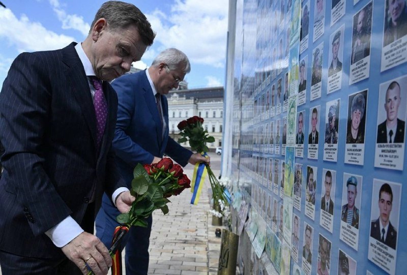 Two men in suits place bouquets of red and yellow roses at the base of a memorial wall covered with rows of portrait photographs of individuals.