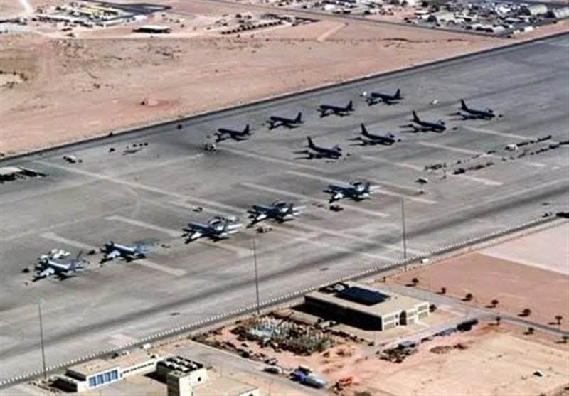 An aerial view shows numerous large military aircraft parked in rows on a desert airfield tarmac, with support buildings visible nearby.