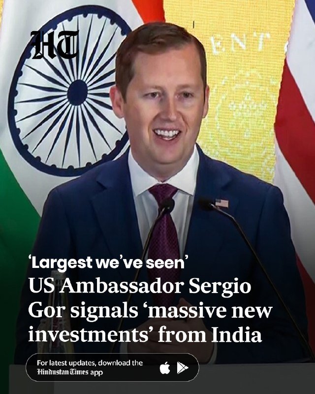 A man in a suit and tie speaks at a podium with microphones, standing before the flags of India and the United States, with overlaid news text.
