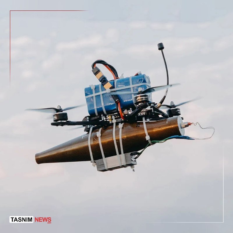 A quadcopter drone carrying a strapped, cone-shaped object with attached wires and blue battery packs hovers mid-air against a pale sky.