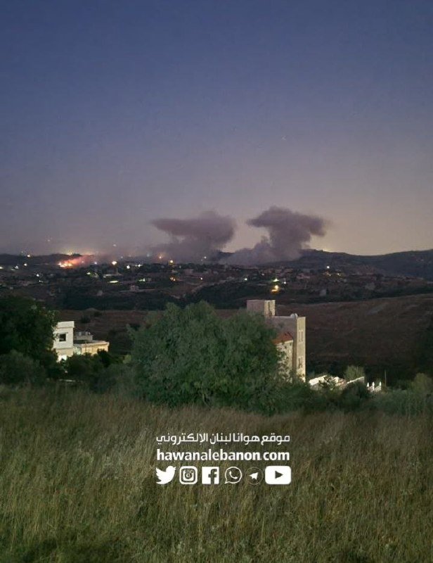Plumes of smoke rise above a hilly landscape dotted with buildings and lights at dusk, with a grassy field and trees in the foreground, overlaid with "hawanalebanon.com" watermarks.