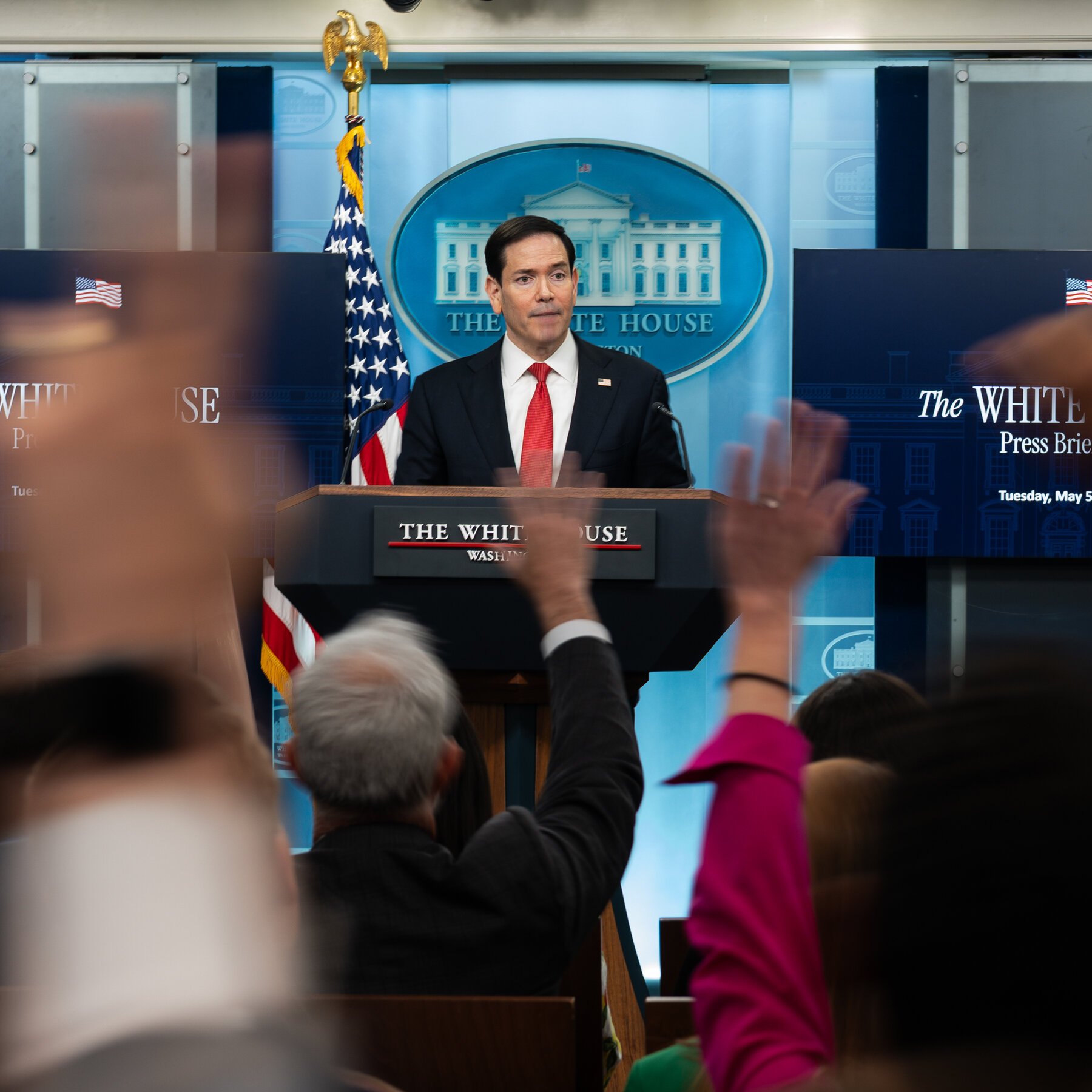 A man in a dark suit and red tie stands at a podium labeled "The White House," facing an audience with raised hands, while screens behind him display "The White House Press Briefing."