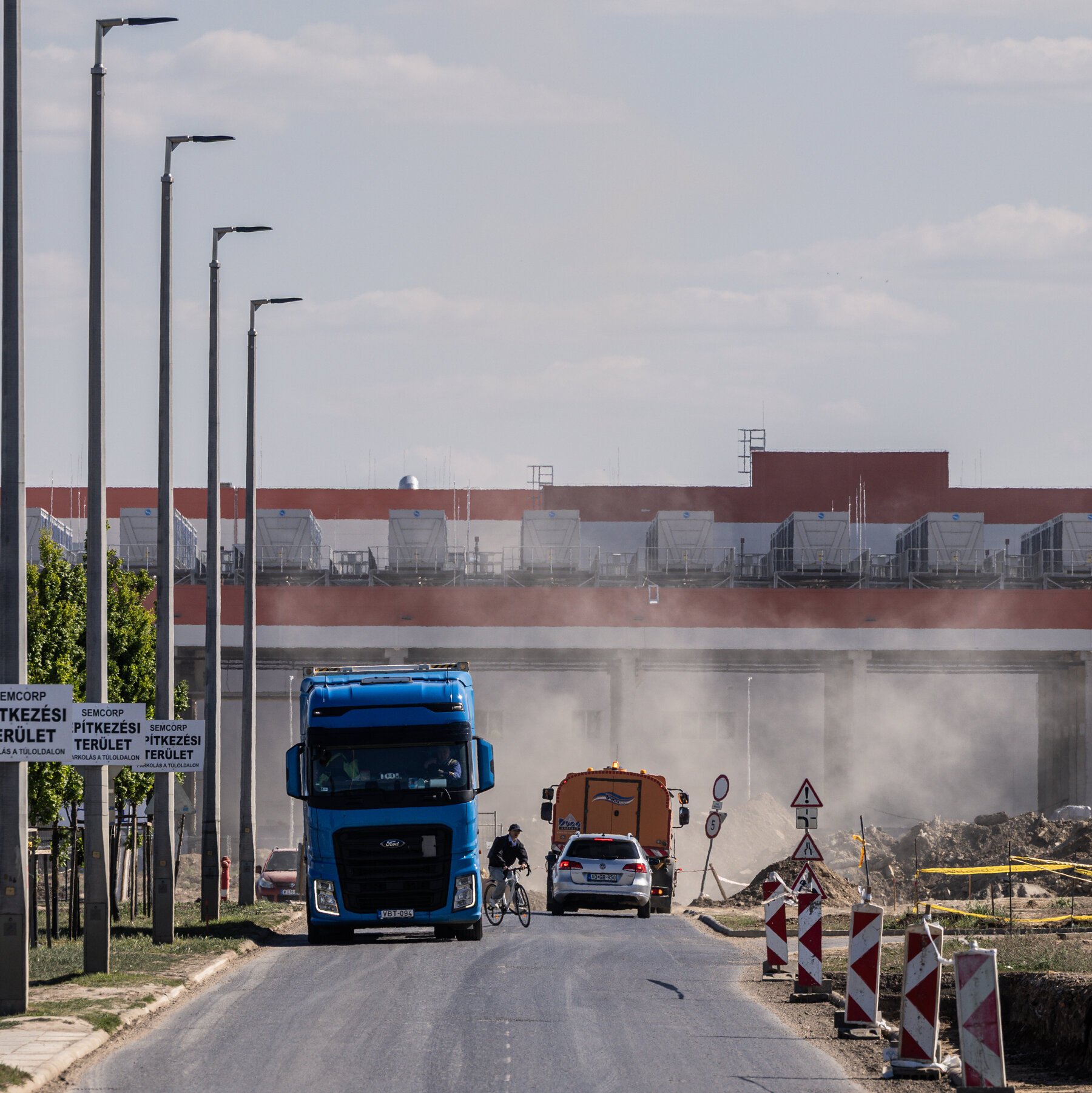 A blue truck, cyclist, car, and orange truck travel down a road past signs and red-and-white barriers, with a large industrial facility visible in the background under a hazy sky.