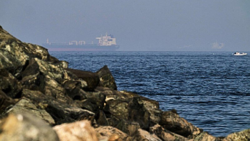 A rocky shoreline in the foreground overlooks a hazy sea, where a large cargo ship and a smaller white boat are visible on the water.