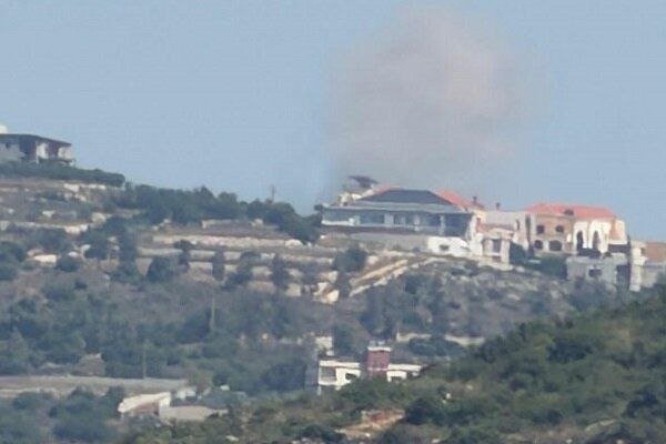 A plume of dark smoke rises from a hillside cluster of houses surrounded by trees and vegetation under a hazy sky.